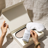 Hands opening a box containing a sleep mask, face mask, and a card with text on a neutral background.